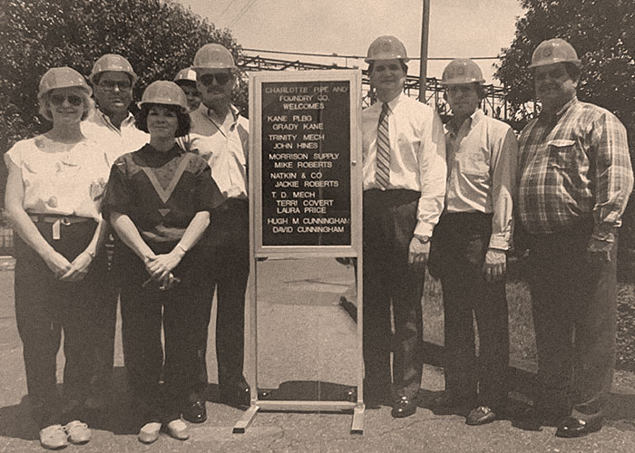 A group of eight people, wearing safety hats, stand beside a signboard with various names listed on it, including Howard Kane Plumbing, posing for a photo outdoors in an industrial area to boost their online presence.