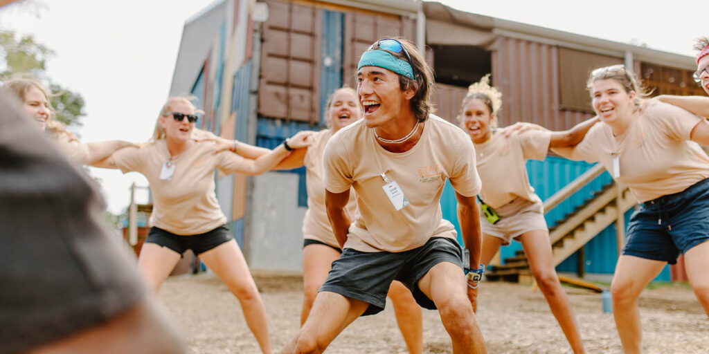 A group of people in matching shirts and shorts engage in an outdoor team activity, smiling and stretching in front of the colorful buildings of Camp Cho-Yeh.